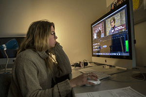 A student sits at a table while looking at a computer screen and holding the mouse, while working on editing a video.