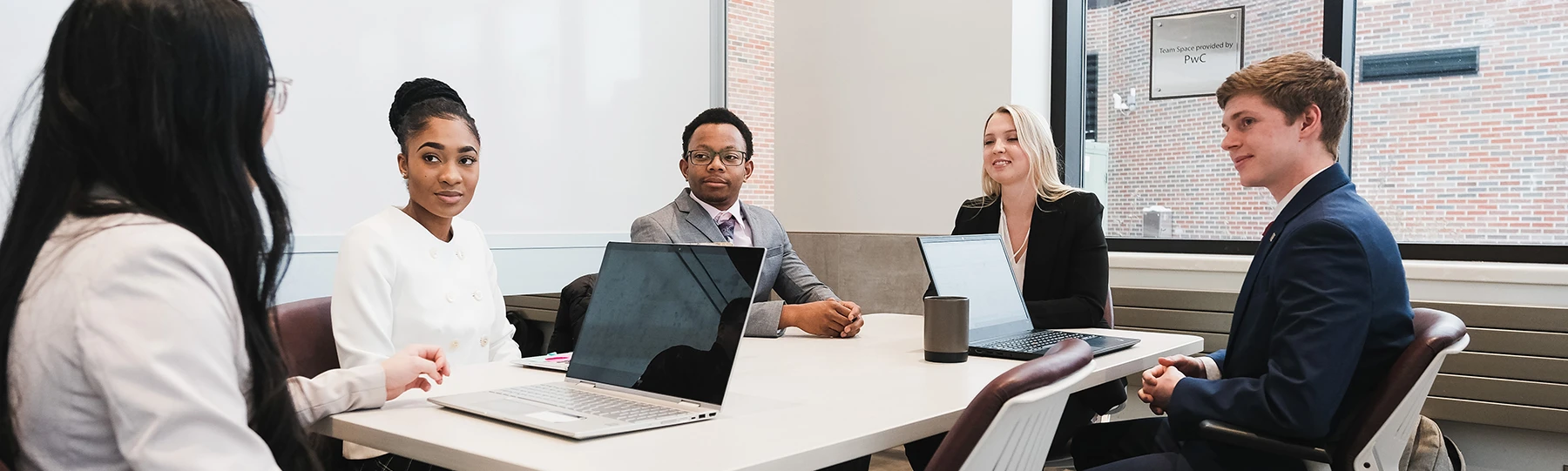 A group of business majors dressed in business attire sit around a long table with laptops open having a discussion with one another.