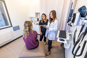 A patient sits on a doctor's office table while two health care providers talk to her.