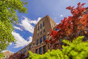 A brick building tower rises into a blue sky with a couple of clouds and sandwiched by budding trees.