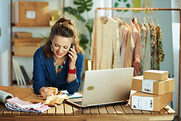 A woman in a blue blouse with brown hair partially pulled back smiles while talking on a phone and leaning against a counter containing folded fabric, boxes, and a laptop.