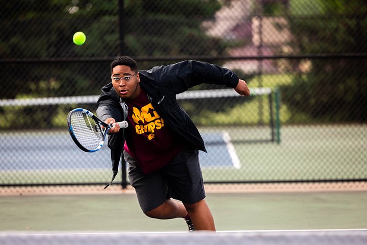 A college student wearing a Fire Up Chips shirt lunges toward a tennis ball while playing tennis.