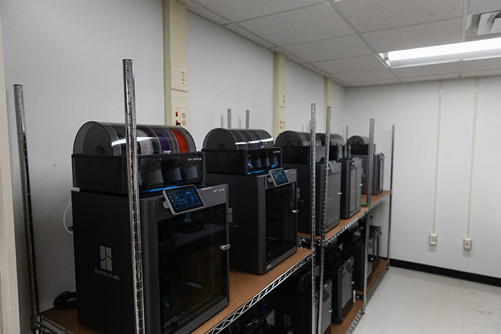 10 3D printers sit on shelves inside a white room with fluorescent lighting.