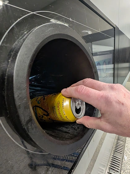 A hand guides an aluminum can into the opening of a bottle and can return machine.