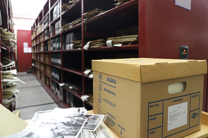 A box of historical exhibits sits on a table in front of a long, red shelving unit filled with other exhibits.