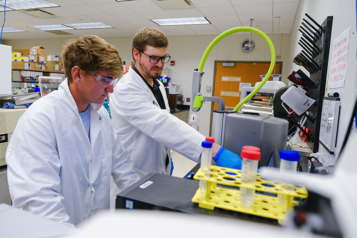 A bearded man with glasses and a white lab coat and blue gloves points at a piece of lab equipment for a young man in safety goggles and a white lab coat.