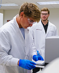 A young man wearing safety goggles, a white lab coat and blue safety gloves looks at lab samples while a bearded man in glasses and a white lab coat looks on.