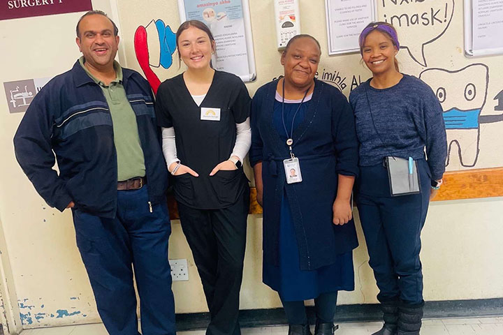 A middle-aged man, a young woman, a middle-aged woman and a younger woman, all wearing blue, stand in a row in front of a bulletin board.
