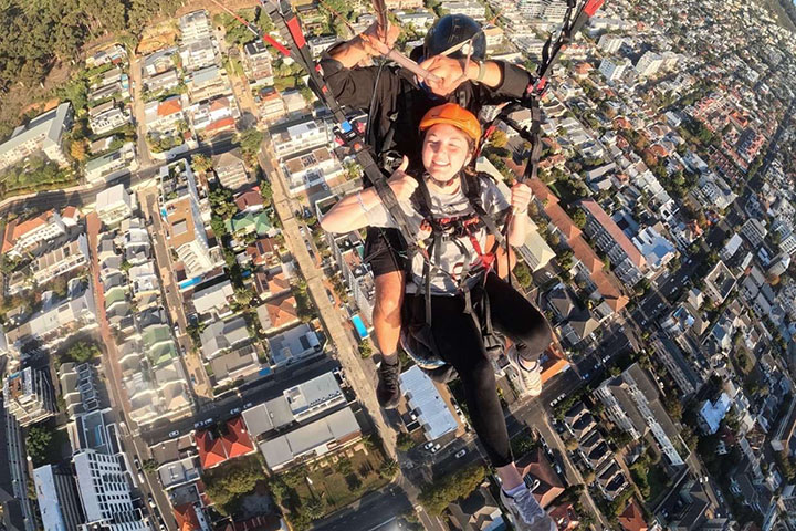 A young woman wearing a helmet and safety harness and tethered to a man wearing a helmet gives a double thumbs up as she parasails against an urban background.
