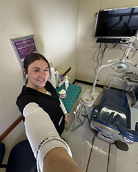 A young woman wearing a long white T-shirt and blue scrubs takes a selfie with dental equipment in the backgound.