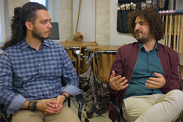 A man with long, dark hair on his head and face and wearing a blue shirt talks to a man with shaggy brown hair wearing a wine-colored jacket and a blue shirt.