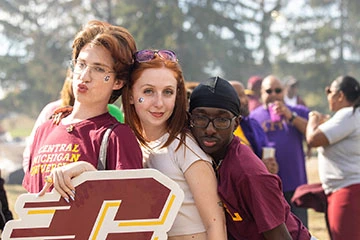 A young man in glasses making a duck face stands in front of a young woman with red hair and a white T-shirt who is standing in front of a young man with a head wrap and glasses, all behind the top of a maroon bold C in italics.