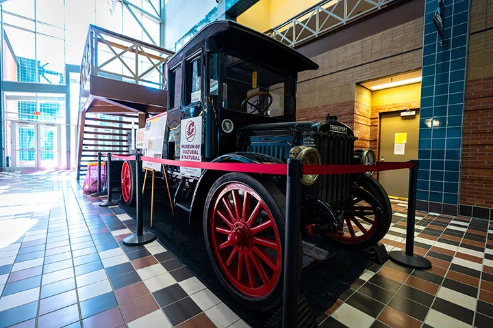 An antique truck painted black with red wheels sits behind a red crowd control tape inside a building.