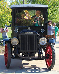 Two people sit in the cab of an antique truck that is outside on a sunny day.