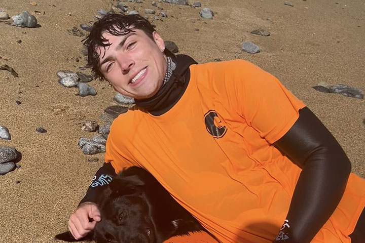 A young man wearing an orange T-shirt and black bodysuit smiles as he lays across a rocky, sandy beach.