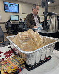 A turkey in a roasting bag sits in a roasting pan on a table as a man in a brown jacket and gray hair talks to a class.