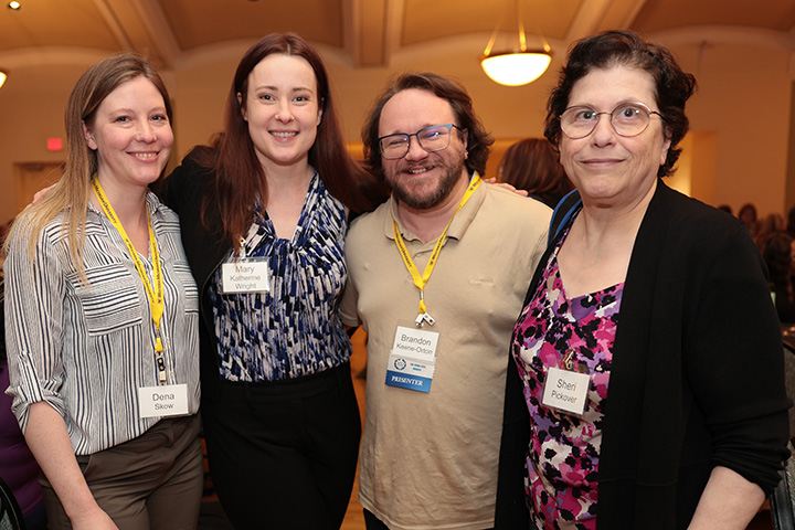Three women and one man pose for a group photo. All four have name tags hanging around their necks.