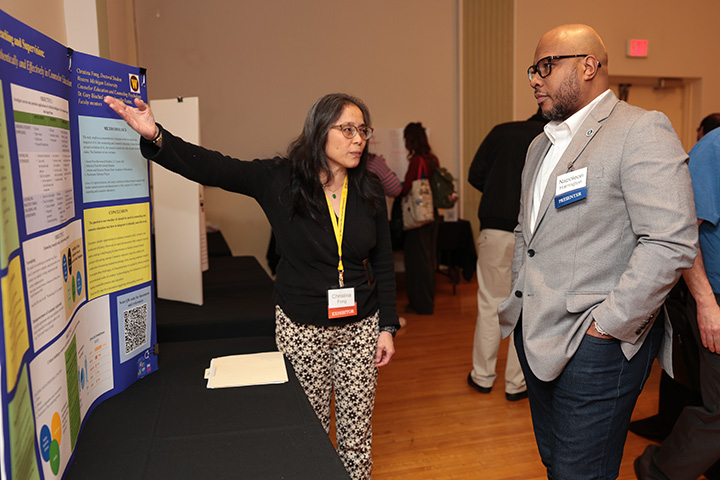 A man listens intently to a woman explaining research shown on a poster board.