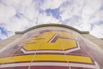 A view looking up at a large CMU action C logo on the side of a building with blue sky and clouds overhead.