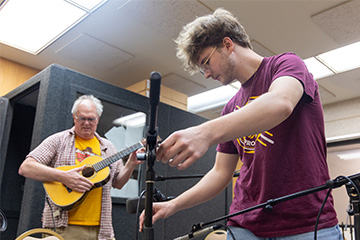 A man playing guitar with another man setting up recording equipment.