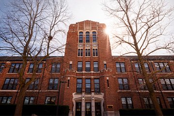 A tall brick building with trees in front of it and sun shining from behind it.