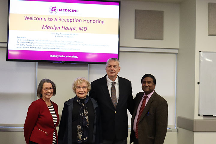 A group of four people wearing suits stand in front of a large display screen.