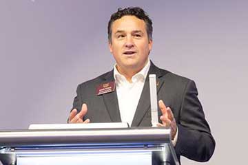 A man in a grey suit stands at a lectern and speaks.