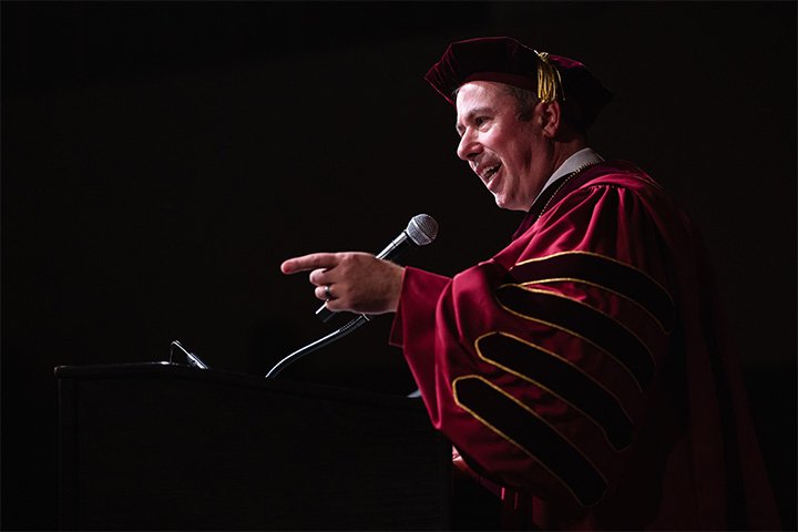 a man in a graduation gown pointing as he speaks