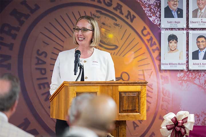A blond woman in a white suit jacket speaks at a podium in front of a mural