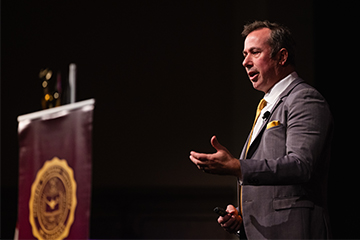 A man in a suit with a yellow tie speaking.