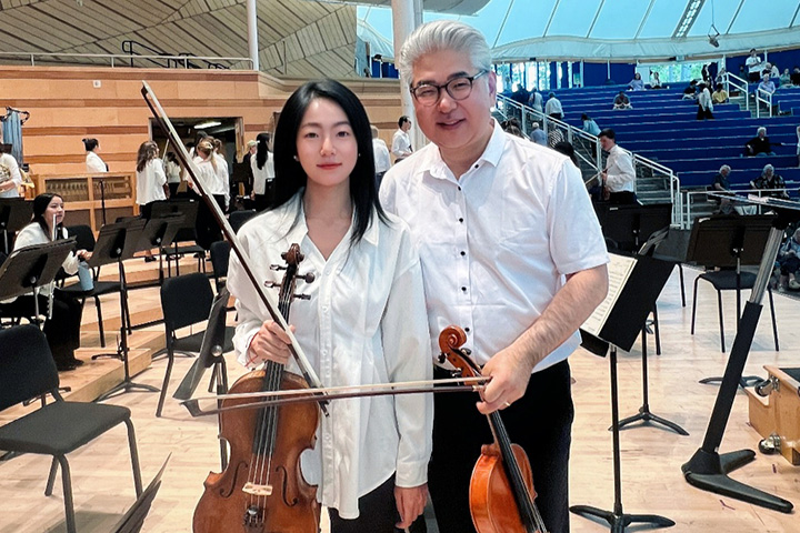 A woman with long black hair and wearing a white button-down shirt holds a violin while a man in glasses with combed-back white hair and also wearing a button-down white shirt stands next to her on a stage amid orchestra furniture.