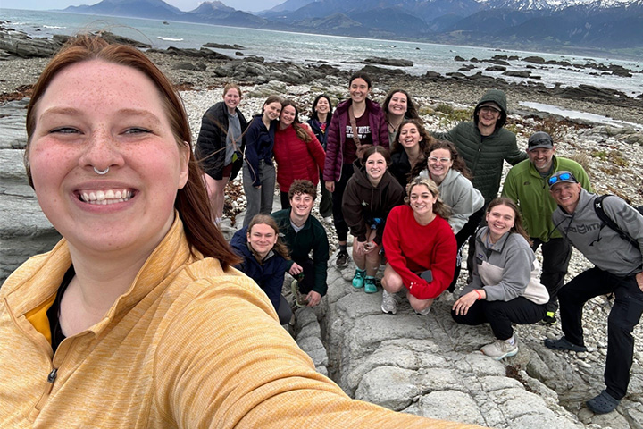 A smiling woman with long brown hair and a yellow top poses for a selfie shot with 16 people in the background standing on rock.