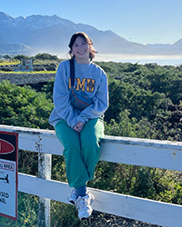 A woman with her hair tied back by a bandana and wearing a gray sweatshirt with CMU on it sits on top of a fence with grass a bay and a mountain in the background.