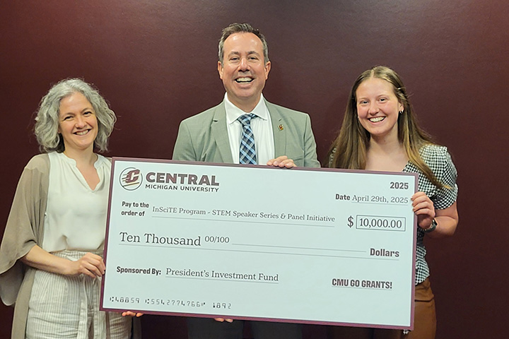 A woman and a smiling man in a suit and a smiling student stand in front of an oversized check with Central Michigan University and Ten Thousand Dollars written on it.