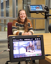 A young smiling woman wearing a sweater with horizontal stripes is recorded on a video camera the screen for which is in the foreground