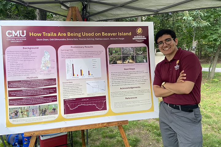 A smiling young man in glasses stands with his arms folded over his maroon polo shirt while standing next to a poster entitled CMU How Trails Are Being Used on Beaver Island