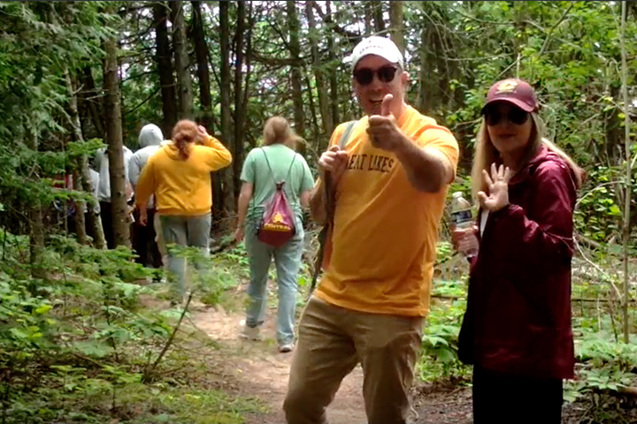 A smiling man in a gold T-shirt and white ball cap gives a thumbs up while standing next to a woman in a maroon top and maroon hat who is waving