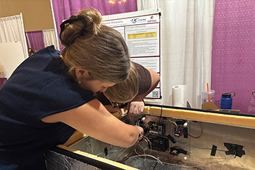 A young woman wearing a blue top and with her hair pulled back into a bun installed electronic circuits while wearing dark nitrile gloves