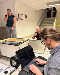 A young man types at a laptop that is connected to the electronic circuits of a wheeled box while two young women laugh in the background