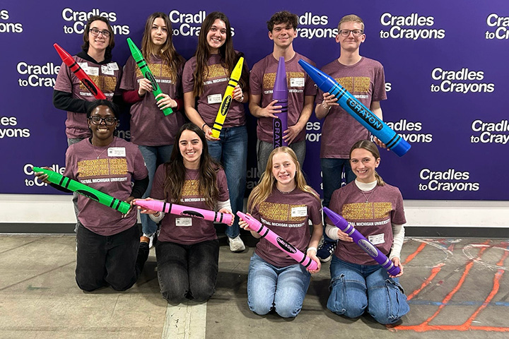 Nine young people wearing maroon and gold t shirts with central michigan university written on them and holding oversized crayons post before a purple backdrop with cradles to crayons written on it multiple times