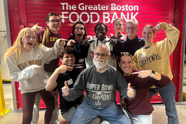 Nine smiling young people wearing sweatshirts of various colors point at a man with white facial hair and giving two thumbs up and wearing a shirt that says super bowl champions stand in front of a red background with the greater boston food bank on it