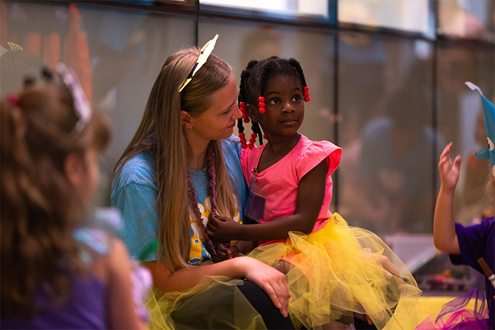 A young girl with briaded hair and a hot pink top and yellow skirt sits in the lap of a woman with brown hair and a blue tee shirt