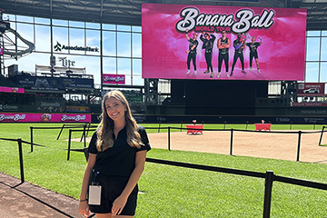 A young woman with long blonde hair parted in the center and wearing a maroon top leans against a railing with a baseball field in the background and a large sign that reads Banana Ball.