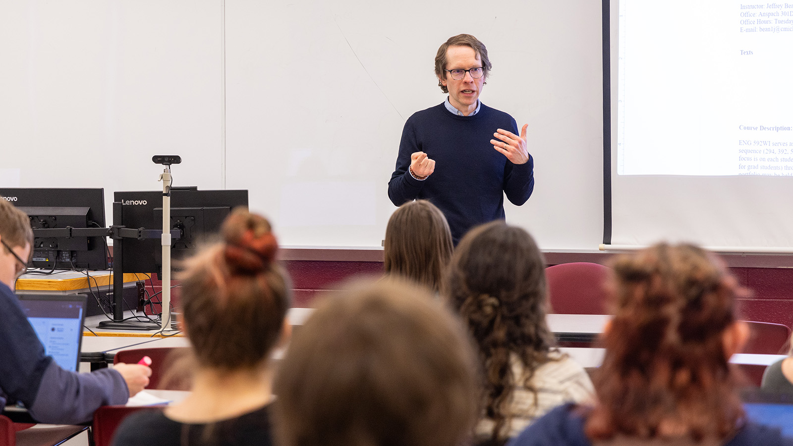 A professor in a black sweater standing in front of a classroom full of students.