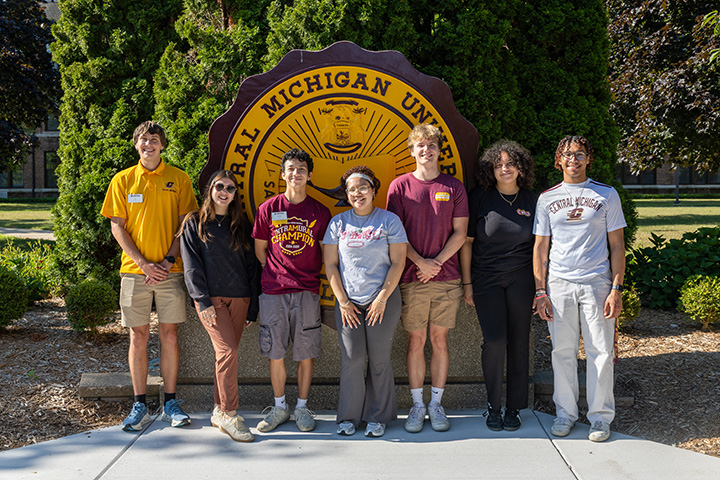 A group of seven CMU student campus ambassadors stand in front of the CMU seal
