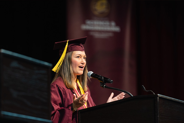 A woman wearing a maroon cap and gown speaks into a microphone.