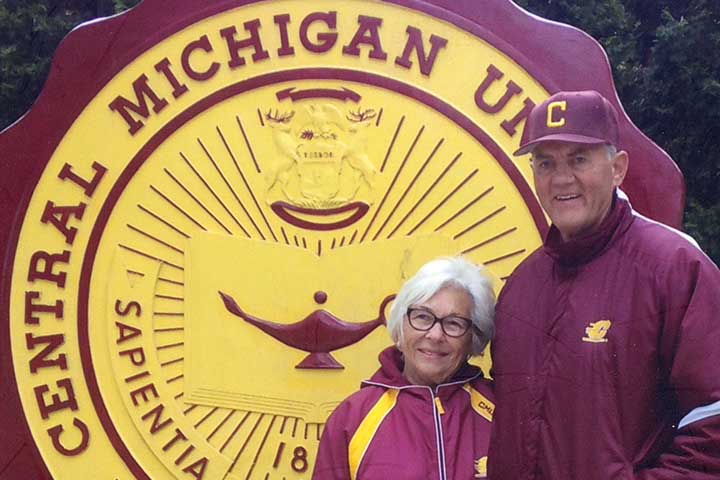 A smiling couple in maroon windbreakers stands in front of the CMU seal on the university campus