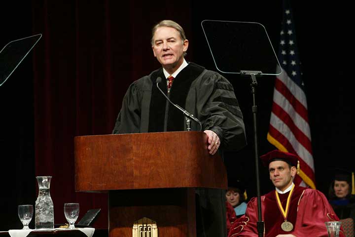 A man in a black commencement robe stands at a podium and speaks into a microphone
