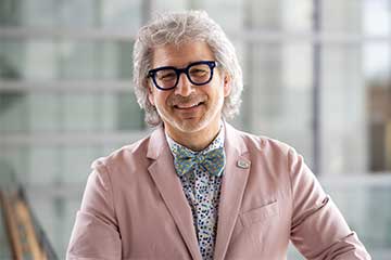 Standing in front of a window, a man with short grey hair wears a pink blazer, light green bowtie and glasses smilies into the camera.