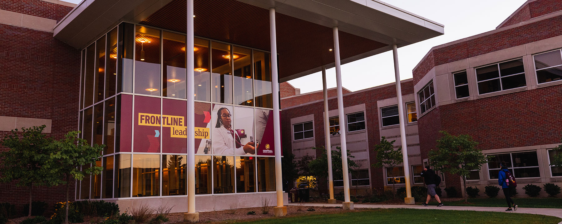 Health Professions building with lights on through glass windows and students walking to building
